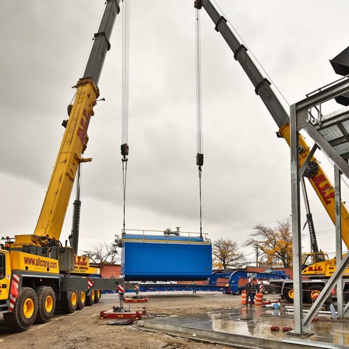 Two cranes lifting a blue boiler unit. NBW Inc.